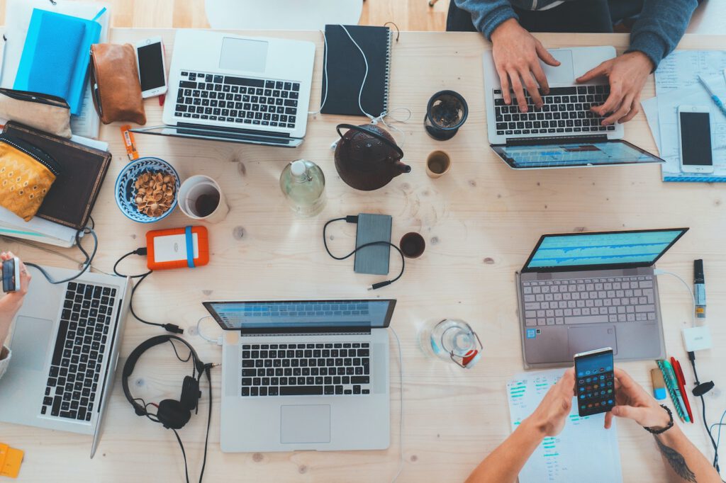 Blick auf einen großen Holztisch an dem mehrere Personen mit Laptops und Handy arbeiten. Kopfhörer, Ladegeräte, Tassen, Kannen und Schalen stehen außerdem auf dem Tisch.